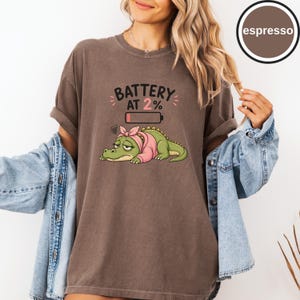 May include: An espresso-coloured t-shirt featuring a graphic of a tired green crocodile wearing a pink headband and sweater. Above the crocodile is the text BATTERY AT 2% with a low battery icon. The shirt has a crew neckline and short sleeves.