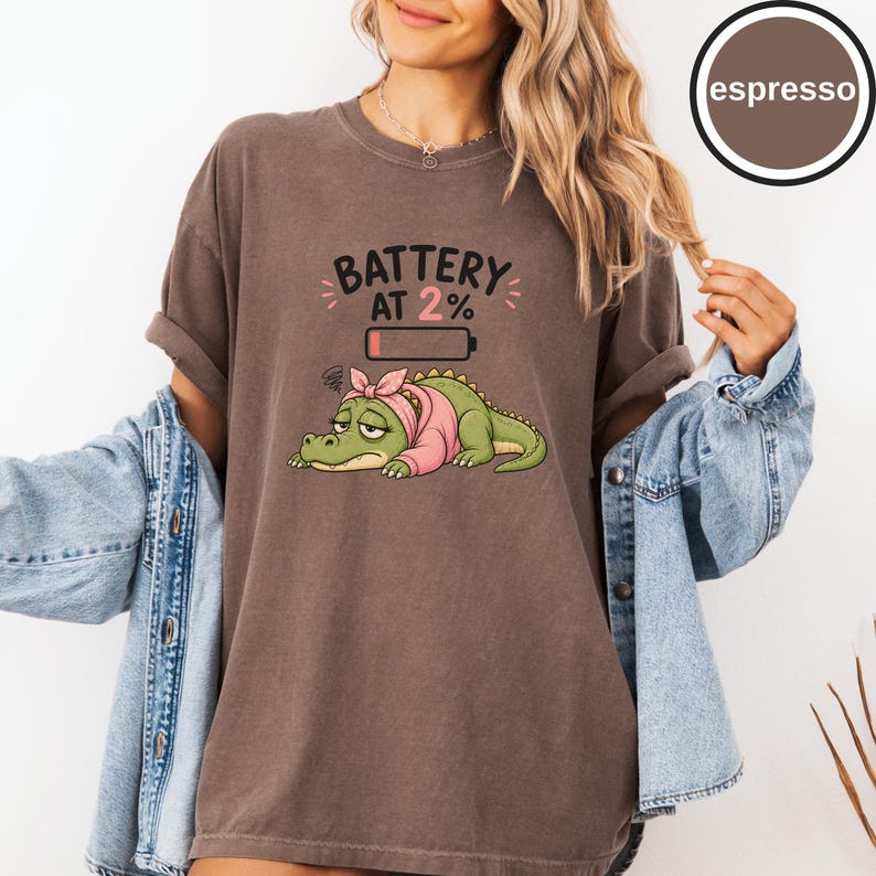 May include: An espresso-coloured t-shirt featuring a graphic of a tired green crocodile wearing a pink headband and sweater. Above the crocodile is the text BATTERY AT 2% with a low battery icon. The shirt has a crew neckline and short sleeves.