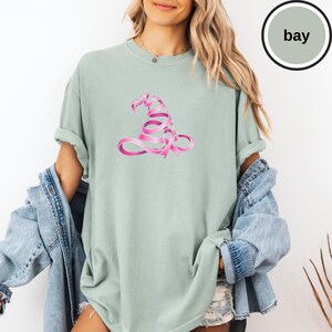 May include: A sage green short-sleeved t-shirt featuring a graphic of a witch hat constructed from a pink awareness ribbon. The shirt has a crew neckline and a loose, relaxed fit. A circular badge in the top right corner contains the text bay.