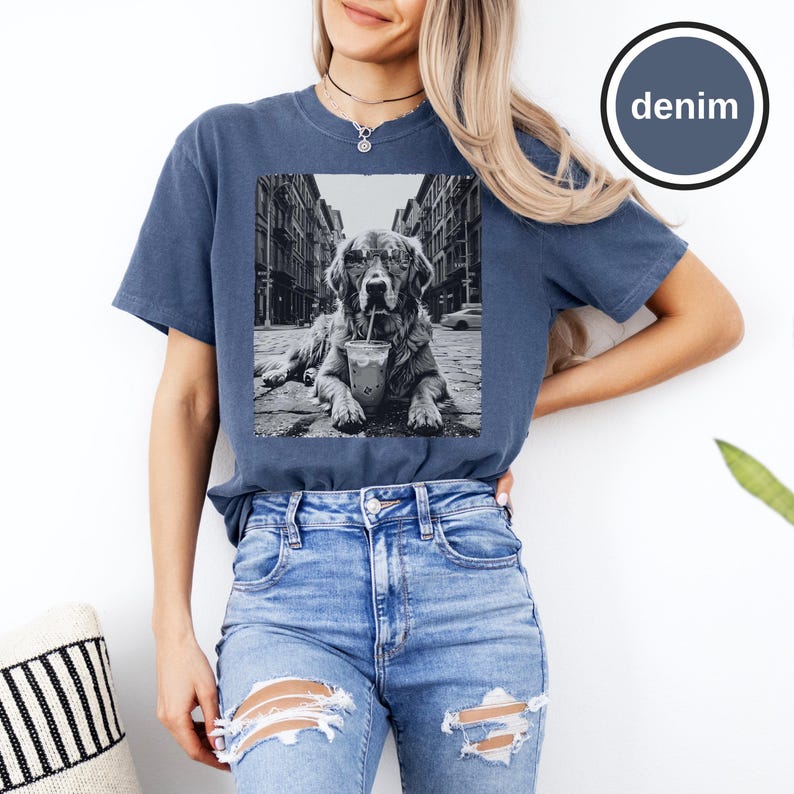 Peut inclure: Un t-shirt bleu denim &agrave; manches courtes avec une impression graphique en noir et blanc repr&eacute;sentant un chien portant des lunettes de soleil et buvant dans un gobelet avec une paille, sur fond de rue urbaine. Col rond et coupe d&eacute;contract&eacute;e.