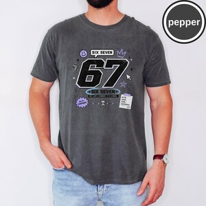 Pu&ograve; includere: Una t-shirt a maniche corte grigio antracite con una stampa grafica che presenta il grande numero 67, il testo SIX SEVEN e piccoli elementi decorativi tra cui una faccina sorridente, una corona, stelle e una nota con 67% chaos 33% confidence.