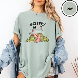 May include: A sage green t-shirt featuring a graphic of a tired green alligator wearing a pink headband and sweater. Above the alligator is a low battery icon and the text BATTERY AT 2%. The shirt has short sleeves and a crew neckline.