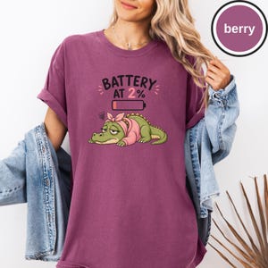 May include: A berry-coloured short-sleeve t-shirt featuring a graphic of a tired green crocodile wearing a pink headscarf and sweater. Above the crocodile is a low battery icon and the text BATTERY AT 2%. The shirt has a crew neckline.