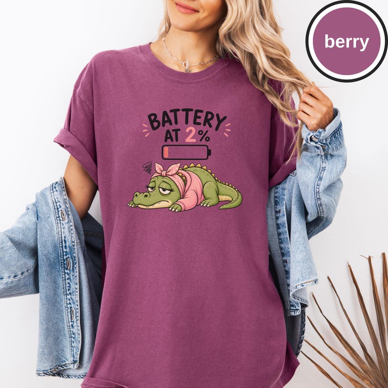 May include: A berry-coloured short-sleeve t-shirt featuring a graphic of a tired green crocodile wearing a pink headscarf and sweater. Above the crocodile is a low battery icon and the text BATTERY AT 2%. The shirt has a crew neckline.