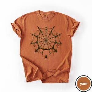 May include: A burnt orange short-sleeve t-shirt featuring a dark green graphic print of a detailed spider web centered on the chest. A small spider hangs from the bottom of the web. The shirt has a crew neckline and a relaxed, casual fit.