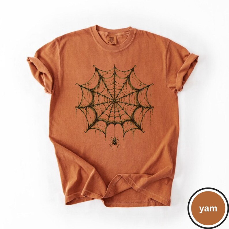 May include: A burnt orange short-sleeve t-shirt featuring a dark green graphic print of a detailed spider web centered on the chest. A small spider hangs from the bottom of the web. The shirt has a crew neckline and a relaxed, casual fit.