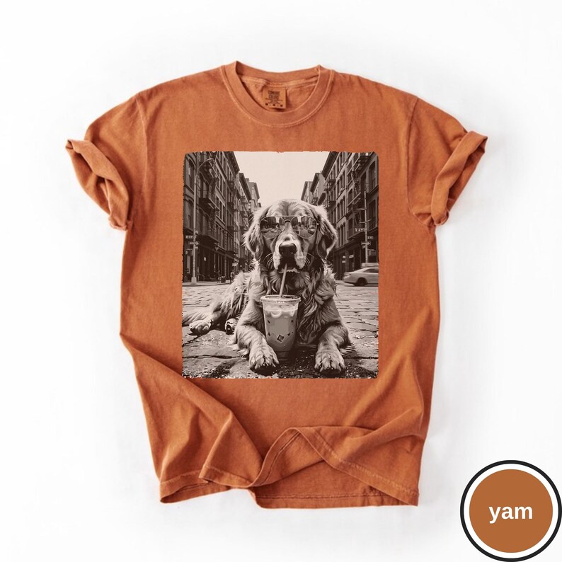 Peut inclure: Un t-shirt &agrave; manches courtes orange br&ucirc;l&eacute; avec une impression graphique centrale en noir et blanc repr&eacute;sentant un golden retriever portant des lunettes de soleil, assis dans une rue avec un gobelet en plastique et une paille devant lui.