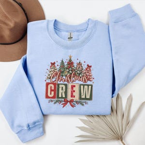 Op de afbeelding: Een lichtblauw sweatshirt met lange mouwen en een grafische print van vijf versierde kerstbomen boven de tekst "Christmas CREW" in rode en groene blokletters, geaccentueerd met kleine sterretjes en een rode strik onderaan het ontwerp.