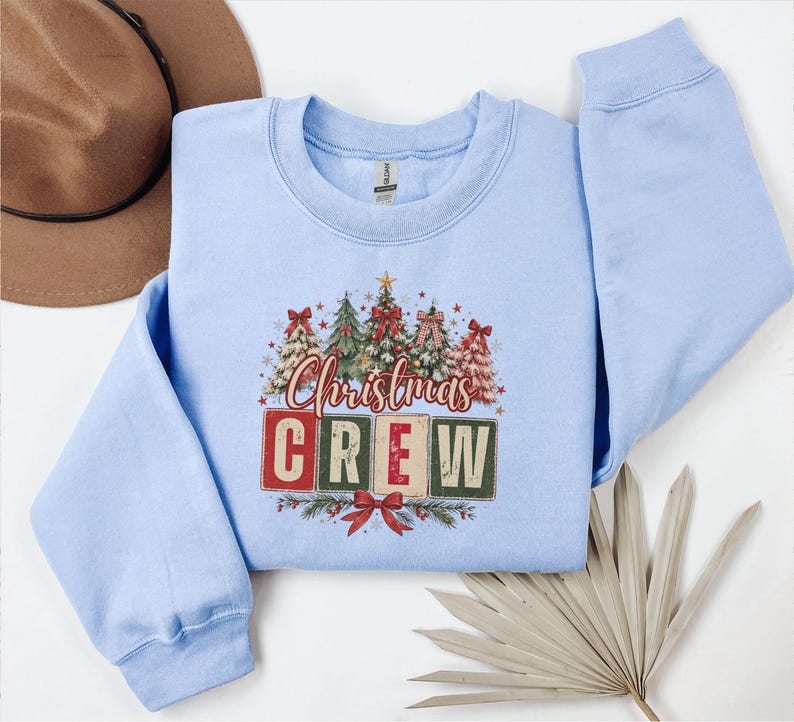 Op de afbeelding: Een lichtblauw sweatshirt met lange mouwen en een grafische print van vijf versierde kerstbomen boven de tekst "Christmas CREW" in rode en groene blokletters, geaccentueerd met kleine sterretjes en een rode strik onderaan het ontwerp.