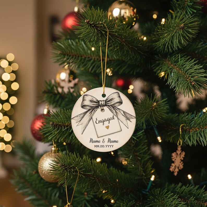 May include: A round white ceramic ornament hanging by a gold string. The design features a black and white illustration of a large bow tied around a square tag with the word Engaged, a small gold heart, and the text Name & Name MM.DD.YYYY.