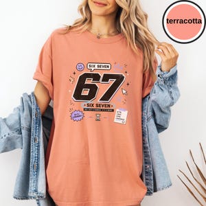 Pu&ograve; includere: Una t-shirt color terracotta con una stampa grafica che presenta i grandi numeri neri 67, il testo SIX SEVEN e varie piccole icone tra cui una faccina sorridente, una clessidra, un cursore del computer e una nota con 67% CHAOS 33% CONFIDENCE.