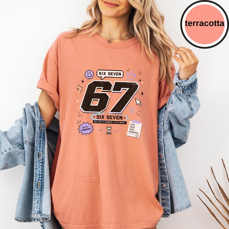 Pu&ograve; includere: Una t-shirt color terracotta con una stampa grafica che presenta i grandi numeri neri 67, il testo SIX SEVEN e varie piccole icone tra cui una faccina sorridente, una clessidra, un cursore del computer e una nota con 67% CHAOS 33% CONFIDENCE.