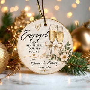 K&ouml;nnte beinhalten: Ein rundes Keramikornament mit zwei Champagnergl&auml;sern, einer beigen Schleife und Blumenmotiven. Der Text lautet Engaged and a beautiful journey begins, Emma & James, und 10.25.2026. Es enth&auml;lt eine goldene Schnur zum Aufh&auml;ngen am Baum.