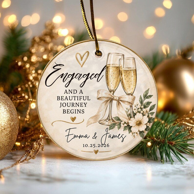 K&ouml;nnte beinhalten: Ein rundes Keramikornament mit zwei Champagnergl&auml;sern, einer beigen Schleife und Blumenmotiven. Der Text lautet Engaged and a beautiful journey begins, Emma & James, und 10.25.2026. Es enth&auml;lt eine goldene Schnur zum Aufh&auml;ngen am Baum.