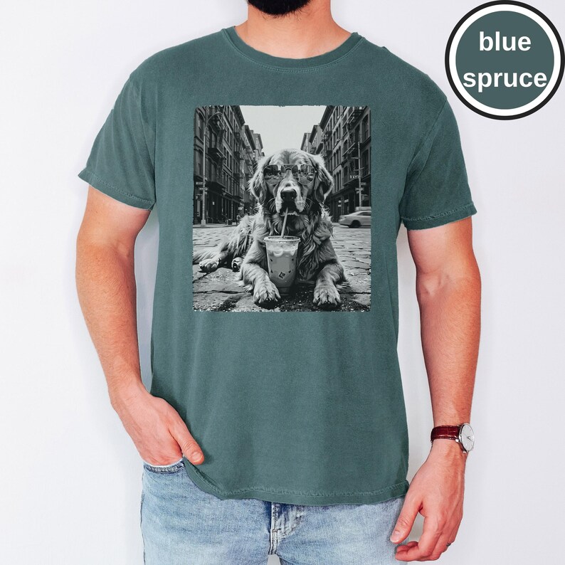Peut inclure: Un t-shirt bleu &eacute;pic&eacute;a avec une impression graphique en noir et blanc repr&eacute;sentant un chien portant des lunettes de soleil et buvant dans un gobelet en plastique avec une paille, allong&eacute; dans une rue. Le t-shirt a un col rond et des manches courtes.
