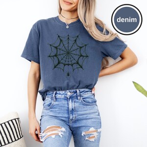 May include: A denim blue short-sleeve t-shirt featuring a dark green spiderweb graphic centered on the chest with a small spider hanging from the bottom of the web. The shirt has a crew neckline and a circular badge in the top right corner with the text denim.