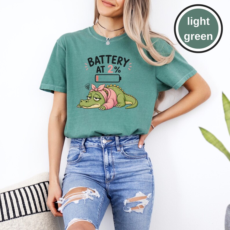 May include: A light green short-sleeve t-shirt featuring a graphic of a tired crocodile wearing a pink headscarf. Above the crocodile, the text reads BATTERY AT 2% with a low battery icon. The shirt has a crew neckline and a relaxed fit.