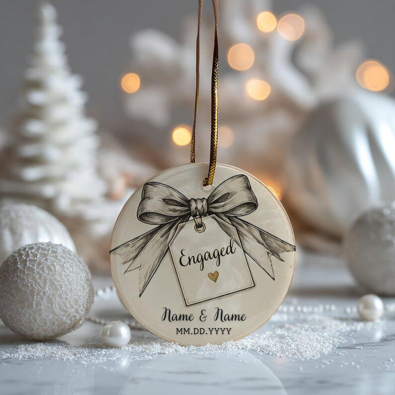 May include: A round ceramic ornament featuring a black and white sketch of a bow tied to a gift tag with the word Engaged and a small gold heart. Below the tag, the text reads Name & Name and MM.DD.YYYY. The ornament hangs from a gold ribbon.