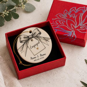 May include: A round white ceramic ornament featuring a black ink illustration of a bow and a gift tag with the text Engaged and a small gold heart. Below the tag, the text reads Name & Name and MM.DD.YYYY. It includes a gold hanging string.