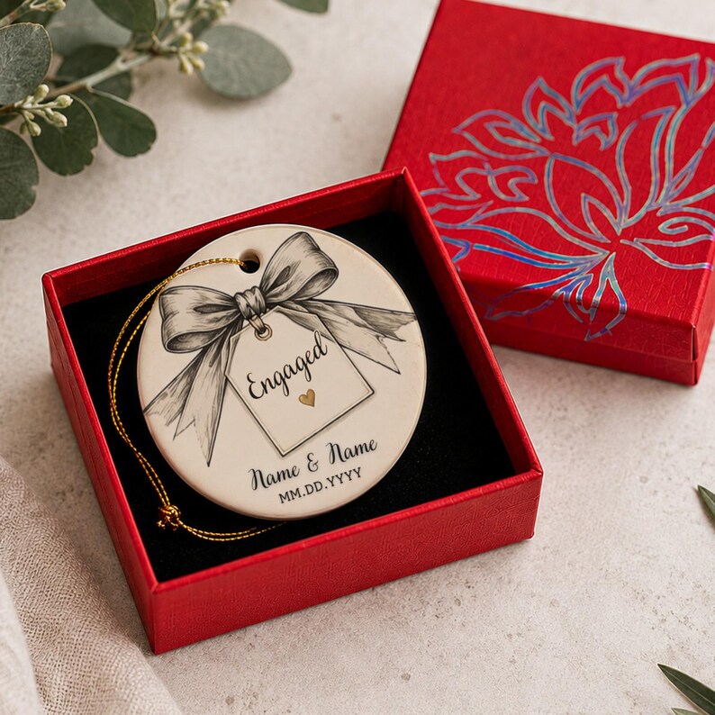 May include: A round white ceramic ornament featuring a black ink illustration of a bow and a gift tag with the text Engaged and a small gold heart. Below the tag, the text reads Name & Name and MM.DD.YYYY. It includes a gold hanging string.