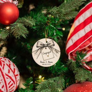 May include: A round white ceramic Christmas ornament featuring a black and white sketch of a bow tied to a gift tag. The tag displays the text Engaged with a small gold heart, followed by the placeholder text Name & Name and MM.DD.YYYY.