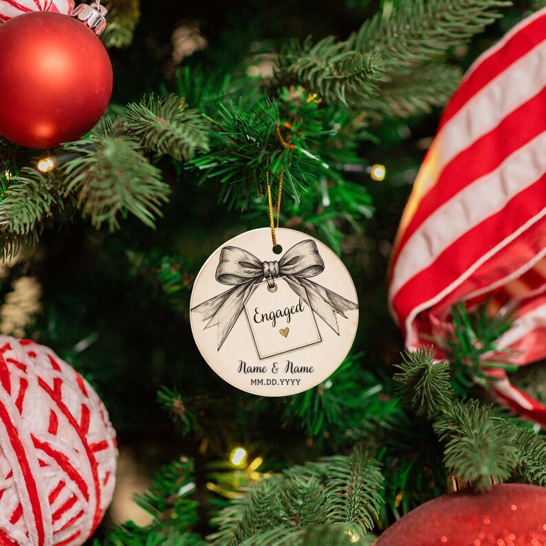 May include: A round white ceramic Christmas ornament featuring a black and white sketch of a bow tied to a gift tag. The tag displays the text Engaged with a small gold heart, followed by the placeholder text Name & Name and MM.DD.YYYY.