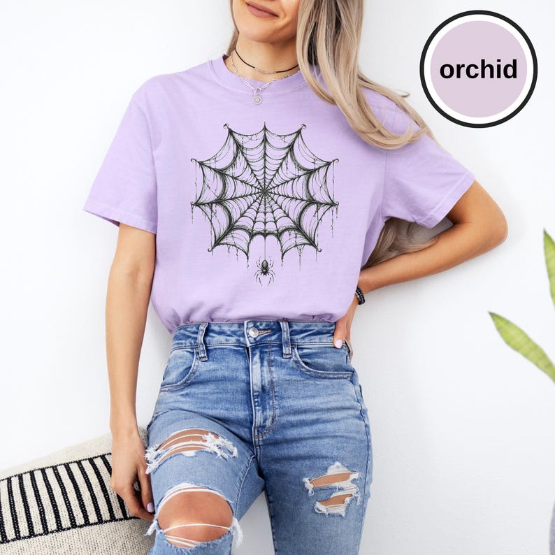 May include: A light purple short-sleeve t-shirt featuring a black ink-style graphic of a spider web with a small spider hanging from the center. The shirt has a crew neckline and is paired with blue denim jeans that have distressed knee holes.