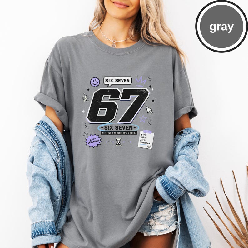 Pu&ograve; includere: Una t-shirt grigia con una grande grafica nera del numero 67 con il testo SIX SEVEN, una faccina sorridente, una corona, una clessidra e una nota che recita 67% CHAOS 33% CONFIDENCE. Il design include anche il testo 6+7=LEGENDARY.