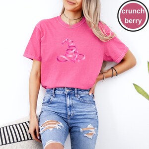 May include: A bright pink short-sleeved t-shirt featuring a graphic of a witch hat formed from a swirling pink ribbon. The shirt has a crew neckline and a relaxed fit. A circular badge in the top right corner displays the text crunch berry.