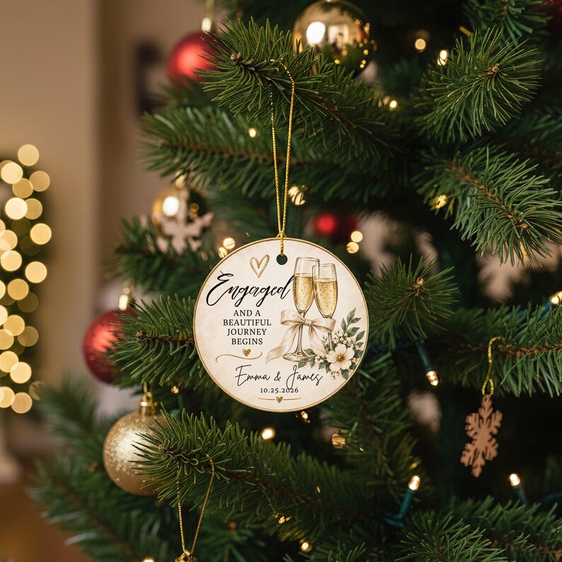 K&ouml;nnte beinhalten: Ein runder, cremefarbener Weihnachtsbaumschmuck aus Keramik, der an einer goldenen Schnur h&auml;ngt. Er zeigt eine Illustration von zwei Champagnergl&auml;sern mit Schleife, Blumenmotiven und dem Text: Engaged and a beautiful journey begins, Emma & James, 10.25.2026.