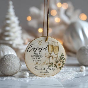 K&ouml;nnte beinhalten: Ein rundes Keramikornament mit zwei Champagnergl&auml;sern, einer Schleife und floralen Akzenten. Der Text lautet: Engaged AND A BEAUTIFUL JOURNEY BEGINS, Emma & James, 10.25.2026. Es enth&auml;lt ein goldenes Aufh&auml;ngeband und kleine goldene Herzmotive.
