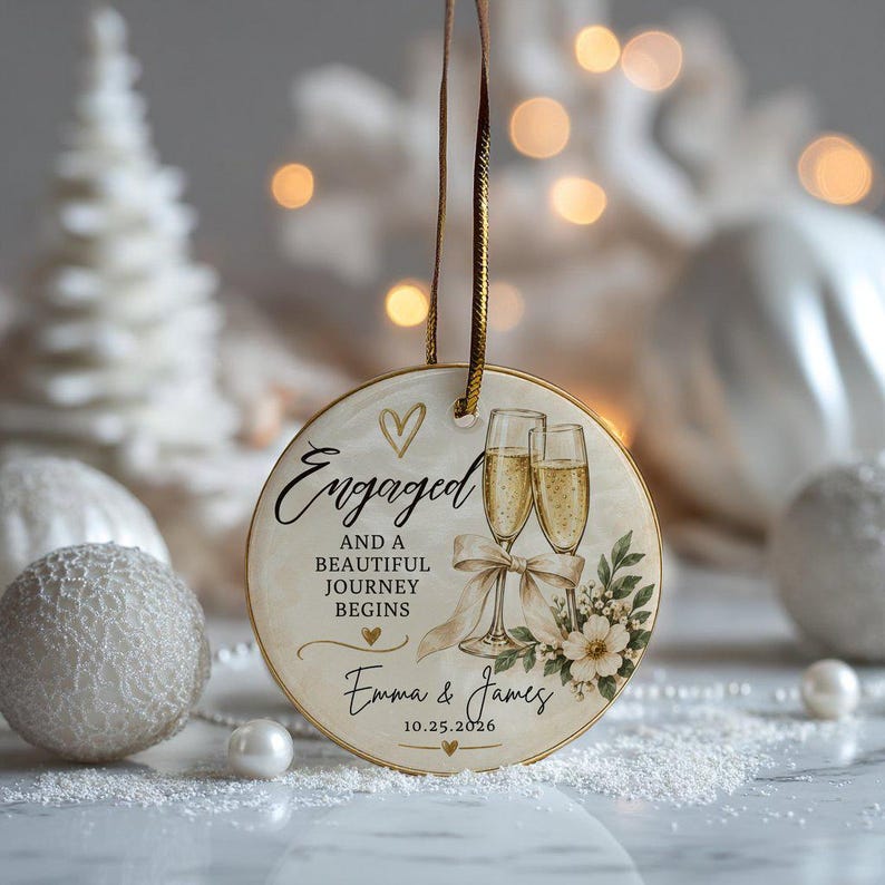 K&ouml;nnte beinhalten: Ein rundes Keramikornament mit zwei Champagnergl&auml;sern, einer Schleife und floralen Akzenten. Der Text lautet: Engaged AND A BEAUTIFUL JOURNEY BEGINS, Emma & James, 10.25.2026. Es enth&auml;lt ein goldenes Aufh&auml;ngeband und kleine goldene Herzmotive.