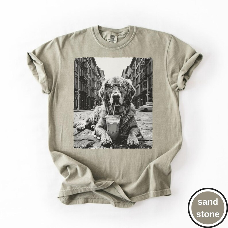 Peut inclure: Un t-shirt couleur sable avec une impression graphique en noir et blanc repr&eacute;sentant un chien portant des lunettes de soleil, assis dans une rue avec un gobelet en plastique et une paille. Le t-shirt a un col rond et des manches courtes.