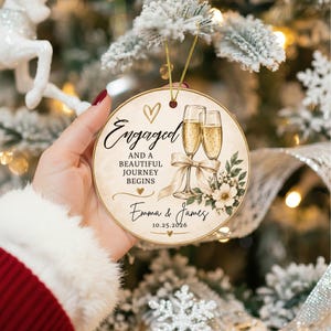 K&ouml;nnte beinhalten: Ein rundes Keramikornament mit zwei Champagnergl&auml;sern, einer beigen Schleife, Blumenakzenten und goldenen Herzen. Der Text lautet: Engaged and a beautiful journey begins, Emma & James, 25.10.2026. Das Ornament h&auml;ngt an einer goldenen Schnur.