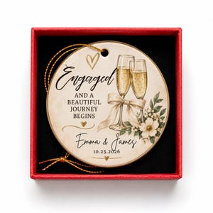 K&ouml;nnte beinhalten: Ein rundes Keramikornament mit goldener Aufh&auml;ngeschnur in einer roten Schachtel. Das Ornament zeigt zwei Champagnergl&auml;ser, eine Schleife und florale Akzente. Der Text lautet: Engaged and a beautiful journey begins, Emma & James, 10.25.2026.