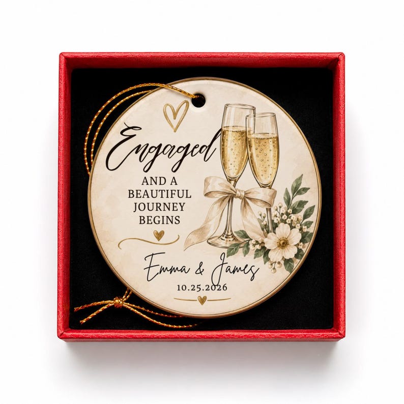 K&ouml;nnte beinhalten: Ein rundes Keramikornament mit goldener Aufh&auml;ngeschnur in einer roten Schachtel. Das Ornament zeigt zwei Champagnergl&auml;ser, eine Schleife und florale Akzente. Der Text lautet: Engaged and a beautiful journey begins, Emma & James, 10.25.2026.