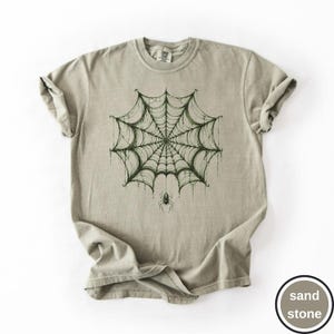 May include: A sand-colored short-sleeve t-shirt featuring a dark green graphic print of a large, intricate spider web centered on the chest. A small spider is depicted hanging from the bottom center of the web. The shirt has a crew neckline.