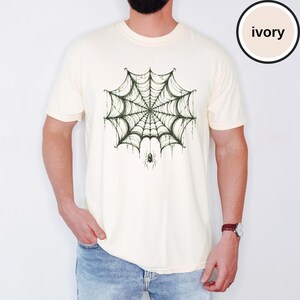 May include: An ivory short-sleeve t-shirt featuring a dark green graphic print of a detailed spider web centered on the chest. A small spider hangs from the bottom of the web. The shirt has a crew neckline and a standard fit, with the color ivory noted.