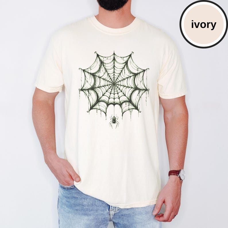 May include: An ivory short-sleeve t-shirt featuring a dark green graphic print of a detailed spider web centered on the chest. A small spider hangs from the bottom of the web. The shirt has a crew neckline and a standard fit, with the color ivory noted.