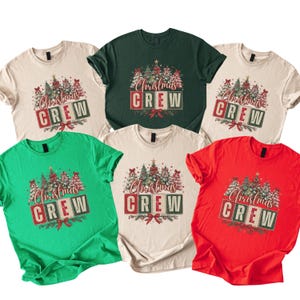 Op de afbeelding: Een collectie van zes t-shirts met korte mouwen in beige, donkergroen, felgroen en rood. Elk shirt heeft een grafische print van kerstbomen en de tekst "Christmas CREW" op blokken met een rode strik aan de onderkant.