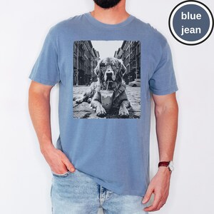 Peut inclure: Un t-shirt bleu jean avec une impression graphique en noir et blanc repr&eacute;sentant un chien portant des lunettes de soleil et buvant dans un gobelet en plastique avec une paille, sur fond de rue urbaine. Le t-shirt a un col rond et des manches courtes.