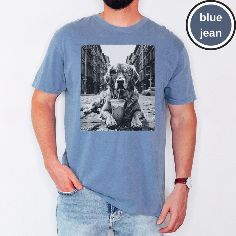 Peut inclure: Un t-shirt bleu jean avec une impression graphique en noir et blanc repr&eacute;sentant un chien portant des lunettes de soleil et buvant dans un gobelet en plastique avec une paille, sur fond de rue urbaine. Le t-shirt a un col rond et des manches courtes.