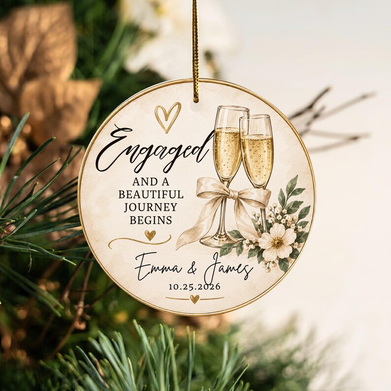 K&ouml;nnte beinhalten: Ein rundes Keramikornament mit goldenem Rand und Aufh&auml;ngeschnur. Es zeigt zwei Champagnergl&auml;ser mit beigem Band, wei&szlig;en Blumen und goldenen Herzen. Der Text lautet: Engaged and a beautiful journey begins, Emma & James, 10.25.2026.