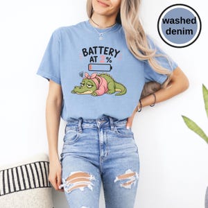 May include: A washed denim blue t-shirt featuring a graphic of a tired green crocodile wearing a pink headscarf and shirt. Above the crocodile is the text BATTERY AT 2% with a low battery icon. The design uses black, pink, and white colours.