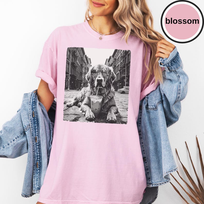 Peut inclure: Un t-shirt rose clair avec une impression graphique en noir et blanc repr&eacute;sentant un chien portant des lunettes de soleil et buvant dans un gobelet en plastique avec une paille dans une rue. Le t-shirt a un col rond et des manches courtes.