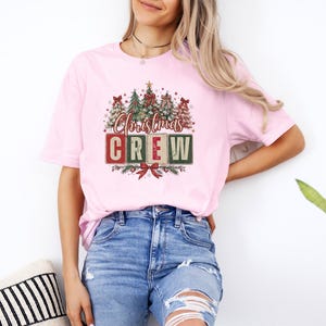 Op de afbeelding: Een lichtroze T-shirt met korte mouwen en een grafische print van verschillende versierde kerstbomen boven de tekst "Christmas CREW" in rode en groene blokletters, geaccentueerd met een rode strik aan de onderkant van het ontwerp.