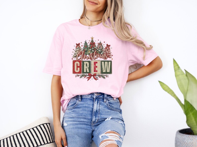 Op de afbeelding: Een lichtroze T-shirt met korte mouwen en een grafische print van verschillende versierde kerstbomen boven de tekst "Christmas CREW" in rode en groene blokletters, geaccentueerd met een rode strik aan de onderkant van het ontwerp.