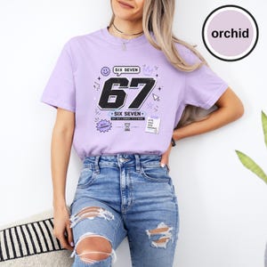 Pu&ograve; includere: Una t-shirt viola chiaro con una grande grafica nera del numero 67, il testo SIX SEVEN, una faccina sorridente, una corona, una clessidra e una piccola etichetta rettangolare con il testo 67% CARDS 33% CONFIDENCE, abbinata a jeans blu strappati.