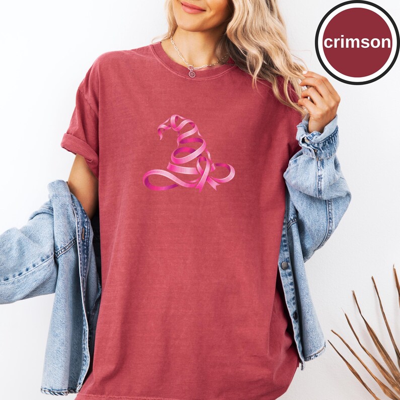 May include: A crimson short-sleeve t-shirt featuring a graphic of a witch hat constructed from a pink ribbon. The shirt has a crew neckline and a relaxed fit. A circular badge in the upper right corner contains the text crimson.
