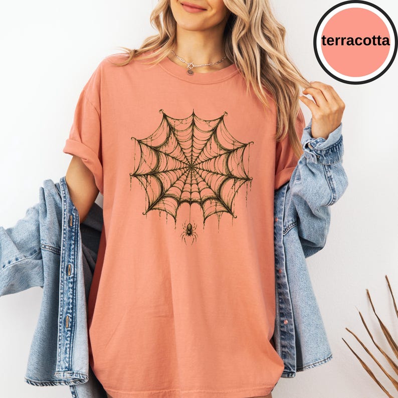May include: A terracotta-colored t-shirt featuring a dark green spiderweb graphic centered on the chest, with a small spider hanging from the bottom of the web. The shirt has short sleeves and a crew neckline, worn with a blue denim jacket.